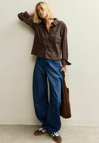 Camicia a quadri marroni con bottoni, jeans blu larghi, sneakers marroni e una borsa marrone. L'outfit presenta una vestibilità comoda e dettagli di design semplici.