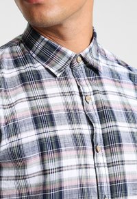 Camisa de manga corta a cuadros con un esquema de colores azul, verde, blanco y rosa, con cuello abotonado y botones de madera.