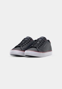 Zapatillas sintéticas negras con cordones grises, suelas blancas y acentos rojos. Forma redondeada con una textura suave y un sutil logo en el lado.