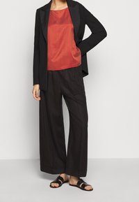 Sort blazer, rød ærmeløs top, løstsiddende sorte bukser og sorte sandaler. Outfittet har et skræddersyet design og en glat overflade.