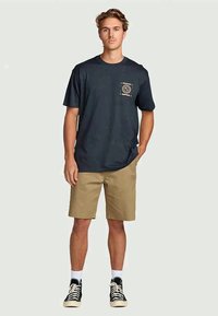 Jonge man staand met zijn handen in zijn zakken, gekleed in een zwart graphic T-shirt, kaki shorts, witte sokken en zwarte sneakers, op een effen achtergrond.