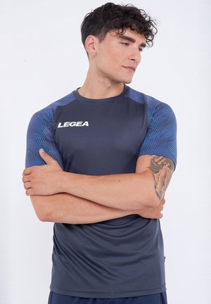 Uomo che indossa una maglietta sportiva LEGEA blu navy con maniche a fantasia, braccia incrociate, tatuaggio visibile sull'avambraccio sinistro, su sfondo bianco uniforme.