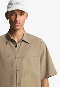 Korte mouw beige shirt met een puntige kraag, twee borstzakken en een gladde textuur. Vergezeld door een witte pet met blauwe tekst.