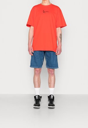 Camiseta roja de gran tamaño con logo negro, pantalones cortos vaqueros y zapatillas negras altas con detalles blancos. La modelo está de pie contra un fondo liso.