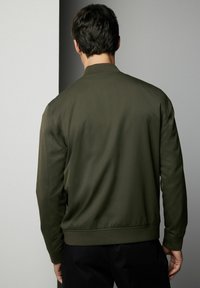 Dunkelgrüne Bomberjacke mit glatter Textur, gerippten Bündchen und Saum. Verfügt über ein einfaches, schmuckloses Design mit einem klassischen Kragen.