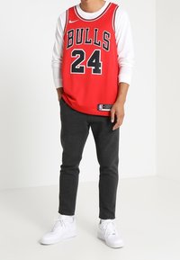 Červená basketbalová dres s nápisem "BULLS" a číslem "24" v černé barvě, vrstvený přes bílou dlouhou tričko. Doplňuje tmavými tepláky a bílými teniskami.
