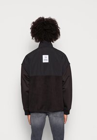 Černá a šedá bunda s vysokým límcem, texturovanou látkou a bílou logovou nášivkou na zádech. Má žebrované manžety a lem.