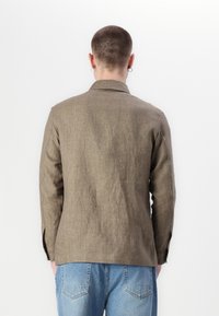 Brown linen blazer with a relaxed fit, notched collar, and side slits. Worn with blue denim jeans, showcasing a casual appearance.