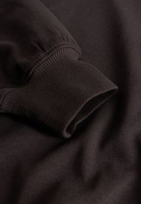 Pruun dressipluus soonikkoes randmetega, mis on siledapinnaline ja lõdva lõikega. Materjal tundub pehme ja kergelt tekstuuriga.