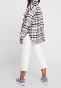 Persoon in een paarse en zwarte geruite lange mouwen shirt, witte cropped jeans en paarse sneakers, staande tegen een witte achtergrond.