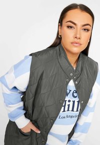 Gilet grigio trapuntato con colletto alto, chiusure a scatto e tasche laterali indossato sopra un maglione a strisce blu chiaro con testo. Texture liscia.