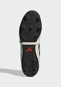 Suela de goma negra con tacos moldeados, parte superior color crema, detalles en franjas plateadas y logo rojo de Adidas en el costado.