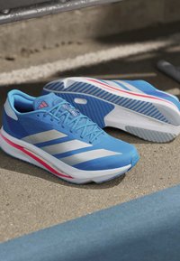 Ζεύγος μπλε παπουτσιών τρεξίματος Adidas με λευκές ρίγες και κόκκινες λεπτομέρειες τοποθετημένα σε μια τσιμεντένια επιφάνεια κοντά σε μια μπλε γραμμή.