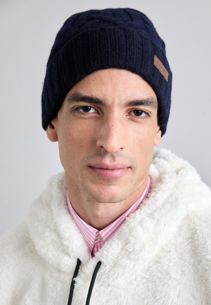 Gorro de malha azul-marinho com um padrão texturizado e um emblema em couro. Usado com um moletom sherpa branco e uma camisa rosa às riscas por baixo.