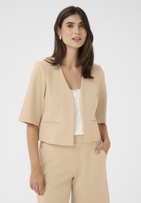 Vaaleanbeige lyhyt blazer, jossa on lyhyet hihat, rakenteellinen muotoilu ja kaksi etutaskua, yhdistettynä valkoiseen puseroon ja yhteensopiviin housuihin.