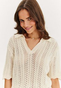 Kremowy sweter w plecionkę z dekoltem w kształcie litery V i krótkimi rękawami, z wzorem ażurowym i ściągaczem pod szyją. Miękka tekstura i przewiewny design.