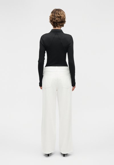 Άτομο με κοντά σγουρά μαλλιά, φοράει εφαρμοστή μαύρη μπλούζα με μακριά μανίκια και άνετο λευκό παντελόνι, εμφανιζόμενο από πίσω με φόντο ένα απλό φόντο.