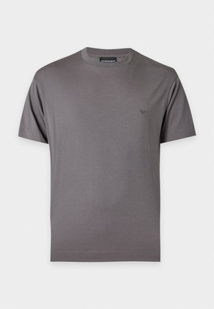 Junger Mann steht, trägt ein dunkelgraues kurzärmeliges Hemd, hellblaue Jeans mit weitem Bein, schwarze Schuhe, mit einer Hand in der Tasche vor weißem Hintergrund.