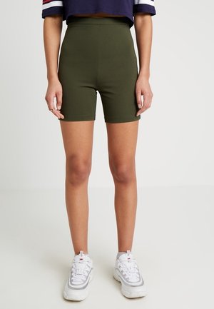 Personne portant un short cycliste taille haute vert olive, un top court à manches courtes bleu marine et des baskets blanches épaisses, debout devant un fond uni.
