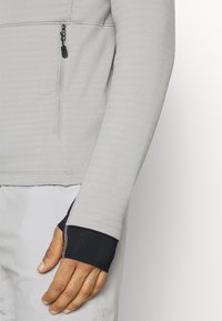 Gros plan sur le bras d’une personne portant une manche longue gris clair avec trou pour le pouce et doublure intérieure noire, à côté d’une poche zippée sur le côté.