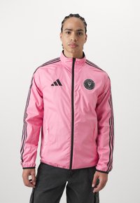 Giacca a vento rosa con strisce nere adidas lungo le maniche. Dotata di una zip frontale e del logo del team sul petto. Materiale liscio e leggero.