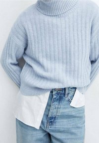 Helle blaue gerippte Rollkragenjacke über einem weißen Hemd mit Kragen, kombiniert mit hellblauen Jeans in hoher Taille. Weiche Textur, lässige Passform.