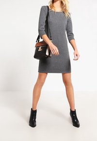 Robe grise texturée avec des manches trois-quarts, décolleté carré et silhouette ajustée. Associée à un petit sac à main noir et beige.