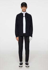 Svart lättviktsjacka med dragkedja framtill, vit skjorta under, svarta träningsbyxor, vita handskar och en svart keps med logotyp. Svarta sneakers med detaljer.