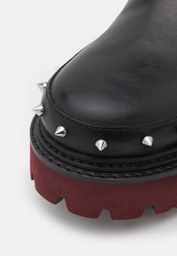 Bout de botte en cuir noir avec des clous argentés en forme de cône et une semelle épaisse texturée rouge foncé sur fond blanc.