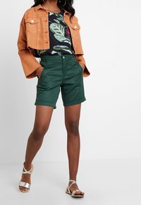 Pomarańczowa dżinsowa kurtka, czarna bluzka w kwiaty, zielone szorty z podwiniętym dołem oraz srebrne sandały na paski zaprezentowane na modelce.