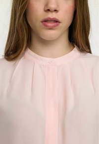 Šviesiai rožinė bluzonė su raukšlėtąja detale, apvaliu iškirpimu ir vienu mygtuko uždarymu priekyje. Pagaminta iš plono, minkšto audinio.