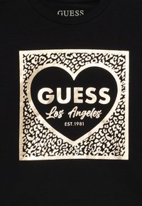 Black fabric with a gold leopard print square containing a gold heart and the text "GUESS Los Angeles EST. 1981" inside.