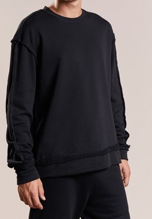 Mężczyzna ubrany w luźną czarną bluzę z postrzępionymi szwami na ramionach, rękawach i dole, w połączeniu z czarnymi spodniami.