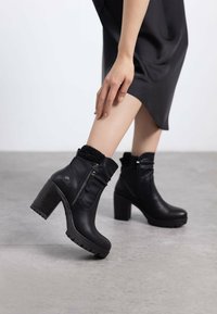 Botas negras hasta el tobillo con un tacón grueso, cremalleras en los lados, tela texturizada en la parte superior y una suela de goma con ranuras de tracción.