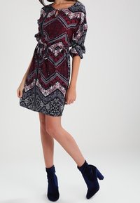 Druckkleid mit einer Mischung aus bordeaux, marineblau und weißen geometrischen Mustern, mit einem Kordelzug in der Taille und einem Knopfdetail am Ausschnitt.