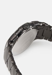 Schwarze Metalluhr mit einem runden Gehäuse, strukturierten Bandgliedern und einer auffälligen Krone an der Seite. Glatte Oberfläche mit einer erhabenen Struktur auf der Rückseite.