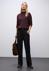 Kvinde iført en blommefarvet turtleneck sweater, sorte vidde bukser, brune ruskinds støvler, som holder en stor brun ruskinds taske mod en ensfarvet baggrund.