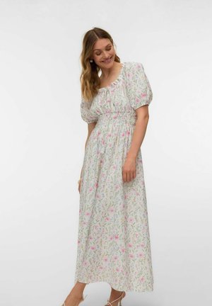 Femme portant une robe maxi blanche à fleurs avec des manches bouffantes et une taille smockée, souriante et regardant vers le bas sur sa gauche.