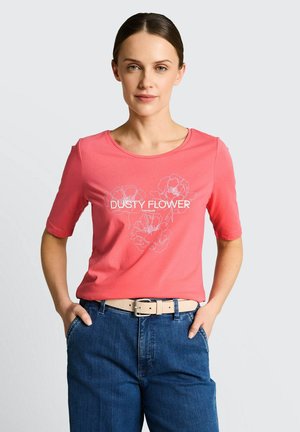 Kobieta o ciemnych włosach w różowej koszuli z napisem „DUSTY FLOWER” i kwiatowym wzorem, wsuniętej w niebieskie dżinsy z beżowym paskiem, ręce w kieszeniach.