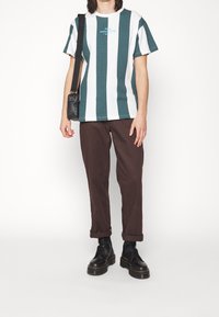 Tričko s krátkým rukávem v tyrkysových a bílých svislých pruzích, s modrým logem. V kombinaci s hnědými kalhotami a černými šněrovacími botami.