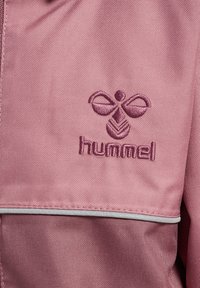 Roze stoffen jas met geborduurd "hummel" logo en een decoratief patroon. Het ontwerp bevat een contrasterende afwerking langs de randen.