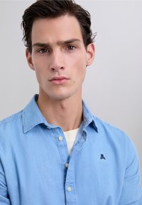 Joven con cabello corto y oscuro, vistiendo una camisa de botones azul claro sobre una camiseta blanca, mirando directamente a la cámara contra un fondo liso.