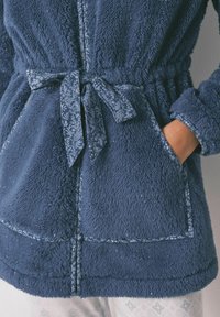 Fluffy marineblauwe robe met een textuur aan de oppervlakte, een gemusterde riem en zakken aan de voorkant. De stof heeft lichte fonkels en decoratieve stiksel.