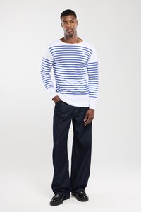 Mand som står iført en hvid langærmet skjorte med blå striber, mørke brede bukser og sorte sko foran en ensfarvet baggrund.