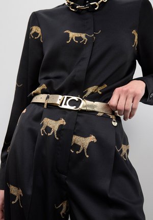 Conjunto negro con estampado de leopardo en dorado, collar de cadena dorada y cinturón metálico dorado con hebilla decorativa, mano sosteniendo el lado del cinturón.