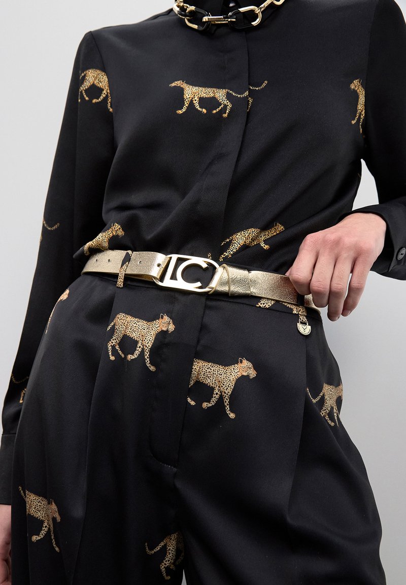 Conjunto negro con estampado de leopardo en dorado, collar de cadena dorada y cinturón metálico dorado con hebilla decorativa, mano sosteniendo el lado del cinturón.