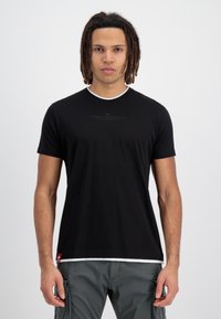 Tricou negru cu maneci scurte, cu guler rotund, cu margine albă și un logo imprimat pe față, asociat cu pantaloni cargo de culoare verde măsliniu.