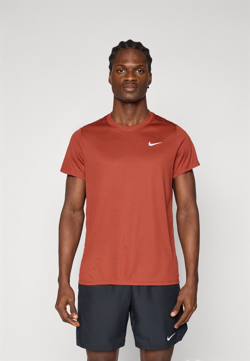 Mann steht frontal und trägt ein rostfarbenes Nike-Shirt und schwarze Nike-Shorts vor einem einfarbigen Hintergrund.