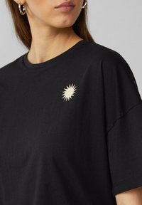 Schwarzes Baumwoll-T-Shirt mit relaxed Fit, das ein strukturiertes, cremefarbenes Sonnenmotiv auf der linken Brustseite aufweist.
