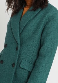 Cappotto di lana teal con design doppiopetto, ampi revers, bottoni neri e dettagli con tasca laterale. Tessuto testurizzato visibile.
